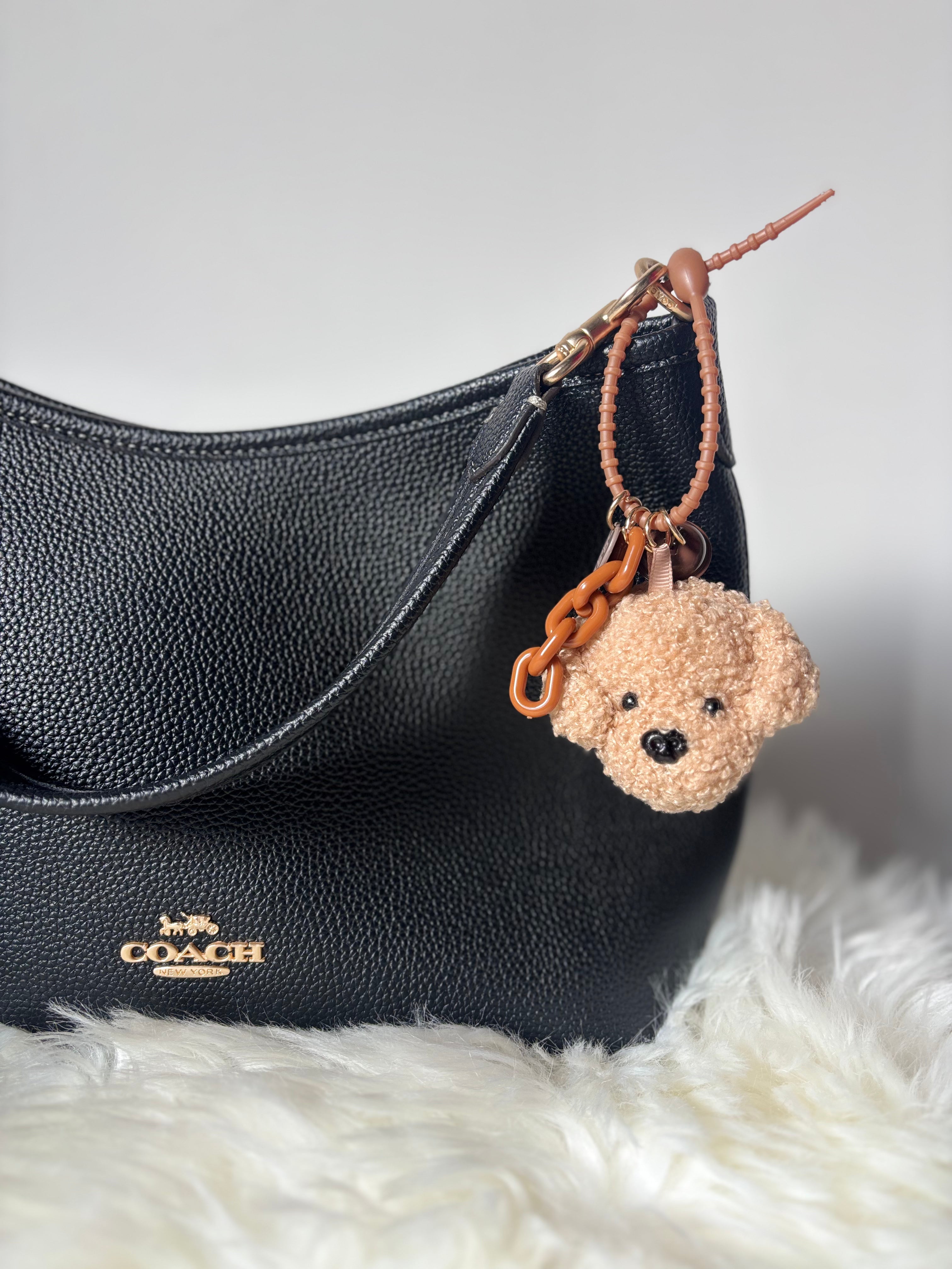 Plush Keychain of Zelci, hanging on COACH bag is displayed on a furry surface. 
